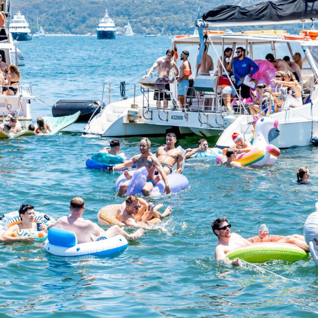 A group of people enjoying a sunny day on a lake, floating on colorful inflatable rings. Several boats are anchored nearby, with more people relaxing and socializing on board. The scene is lively and festive, under a clear blue sky.