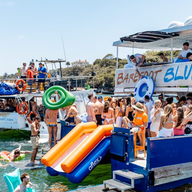 A lively boat party with people in swimsuits enjoying the sun and water. Multiple people are on a double-decker boat with inflatable tubes and slides, surrounded by clear blue water and trees in the background.