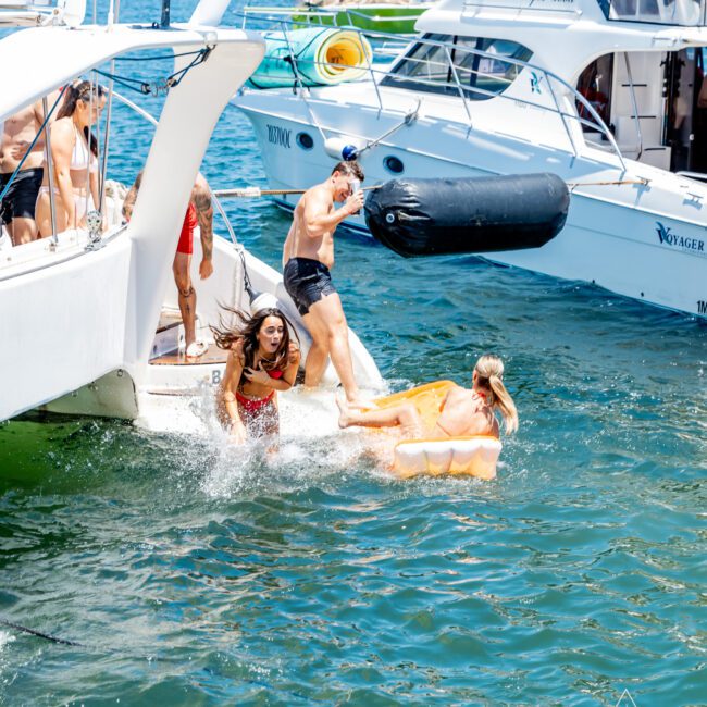 A group of people enjoying a sunny day on a boat in a harbor. Some are stepping into the water, with one person on an inflatable, and others watching. The city skyline is visible in the background. The water is a vibrant blue.