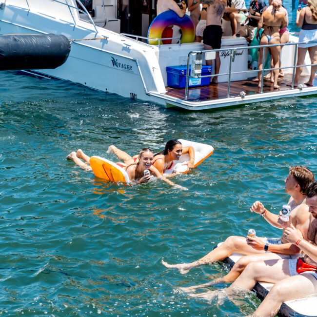 A lively scene on the water with people enjoying a boat party. Two individuals float on inflatables in the water, while others socialize on the boat, holding drinks. The background shows a cityscape under a sunny sky.