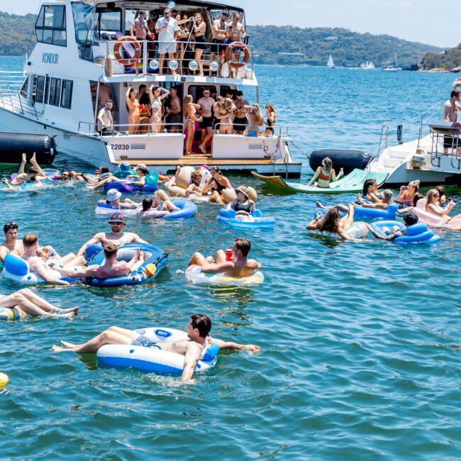 People enjoy a sunny day on the water, lounging on inflatable tubes near a large boat. The boat has a crowd on board, with a scenic backdrop of hills and sailboats in the distance.