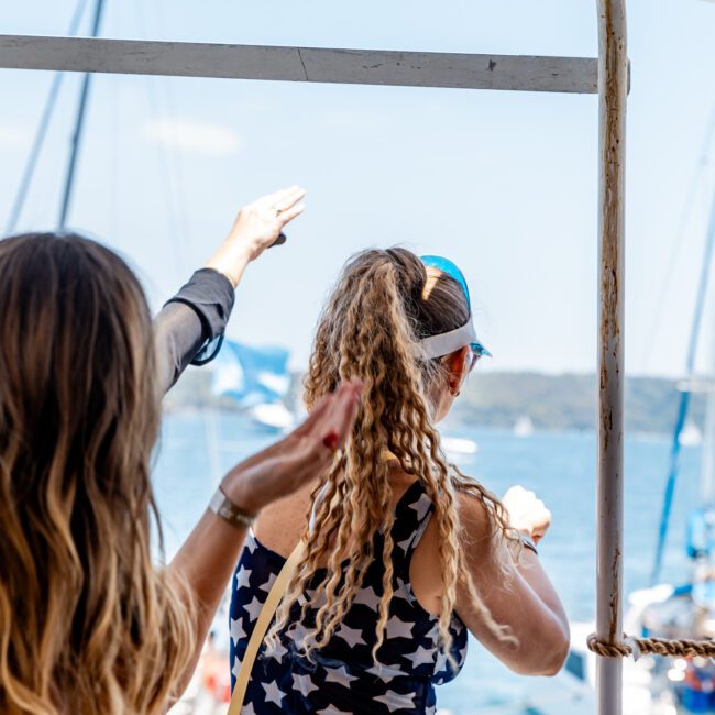 A group of people on a boat joyfully waving at the shore. The woman in the foreground has long wavy hair and is wearing a star-patterned dress and visor. The background features water and blurred sailboats.