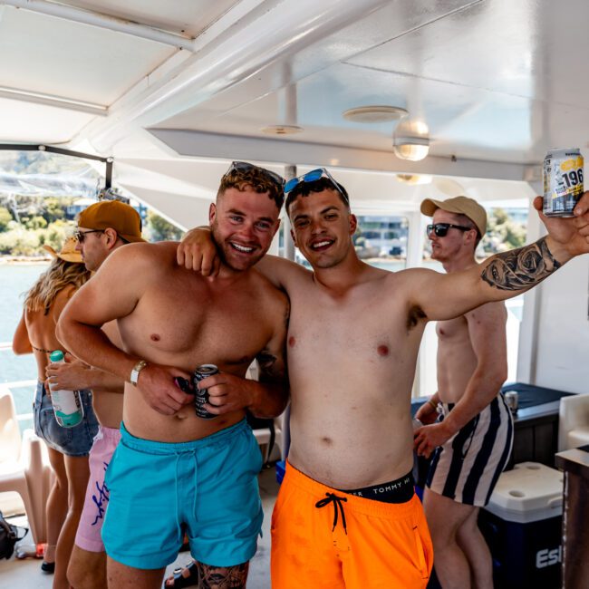 Two shirtless men are smiling and holding drinks on a boat. One wears blue shorts, the other in bright orange. Other people in swimwear are in the background, and a scenic view is visible through the boat's windows.