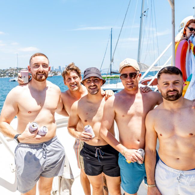 A group of five men in swimwear are standing on a boat, holding drinks and smiling at the camera. The sunny day and clear sky add to the lively atmosphere. A person in a cape is seen in the background.