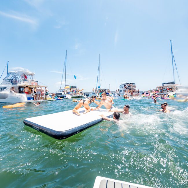 People enjoying a sunny day on the water with floating mats, kayaks, and nearby boats. The scene is lively, with groups swimming and lounging around, set against a clear blue sky with a few yachts anchored nearby.