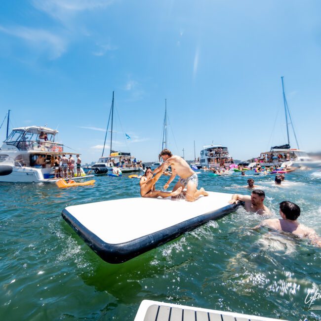 People enjoying a sunny day on the water with boats and inflatables. Some are swimming, while others relax on a large floating mat. The sky is clear, and the atmosphere is lively and fun.