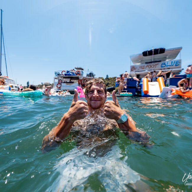 A person in the water gives two thumbs up, smiling with boats and inflatable toys in the background under a clear sky. The scene appears lively and festive with people enjoying a sunny day on the water.