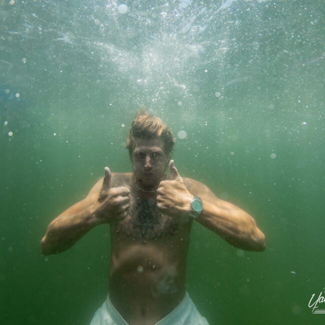 A man underwater gives two thumbs up. He's shirtless, wearing a watch and light-colored shorts. The water around him is greenish, and bubbles rise from his movement, indicating he's submerged.