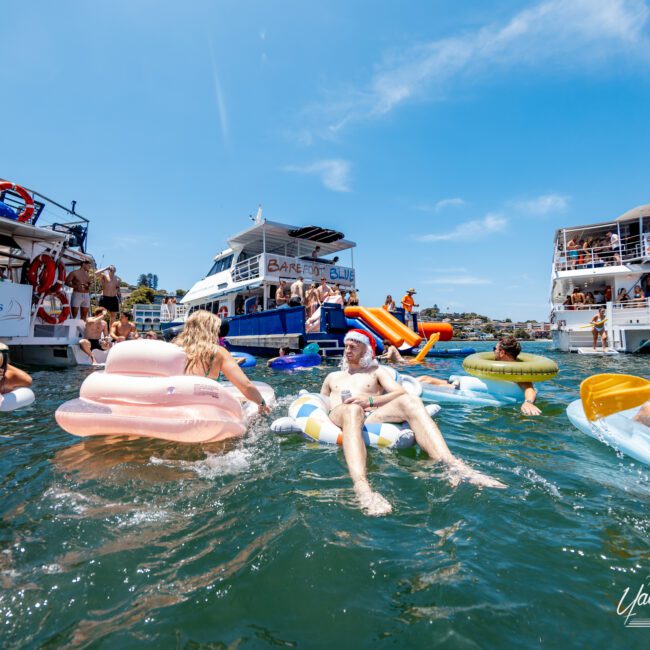 People enjoying a sunny day on a river or lake, lounging on colorful inflatable floats. Several boats are docked nearby, including one with a "Barrel of Fun" sign. The water is clear, and the sky is bright blue.