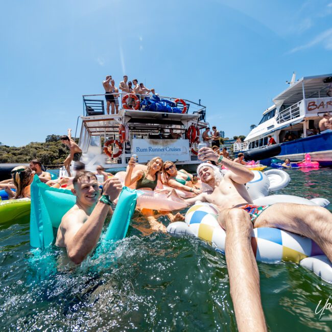 A lively group of people in swimsuits enjoying a sunny day on the water. Some are relaxing on inflatable floats, while others are gathered on a nearby boat. The atmosphere is festive and fun.