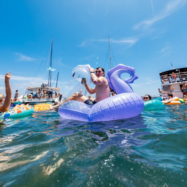 A person in sunglasses is sitting on a purple inflatable dragon float, playfully splashing water with a can in hand. They are surrounded by people and boats on a sunny day, with a clear blue sky above.