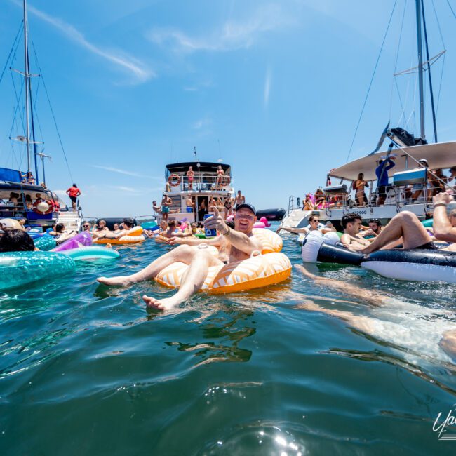 People enjoying a sunny day at a yacht party, floating in the water on colorful inflatable rings. Several yachts are anchored nearby, with more people aboard. The scene is lively and vibrant under a clear blue sky.