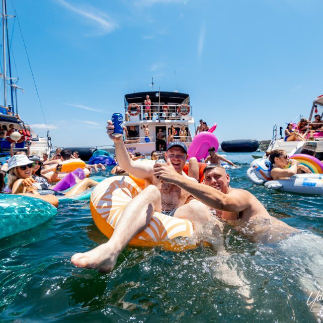 People enjoying a sunny day on a lake, floating on inflatable rafts with drinks. Several boats are anchored nearby. The scene is lively, with individuals relaxing and socializing in the water under a clear blue sky.