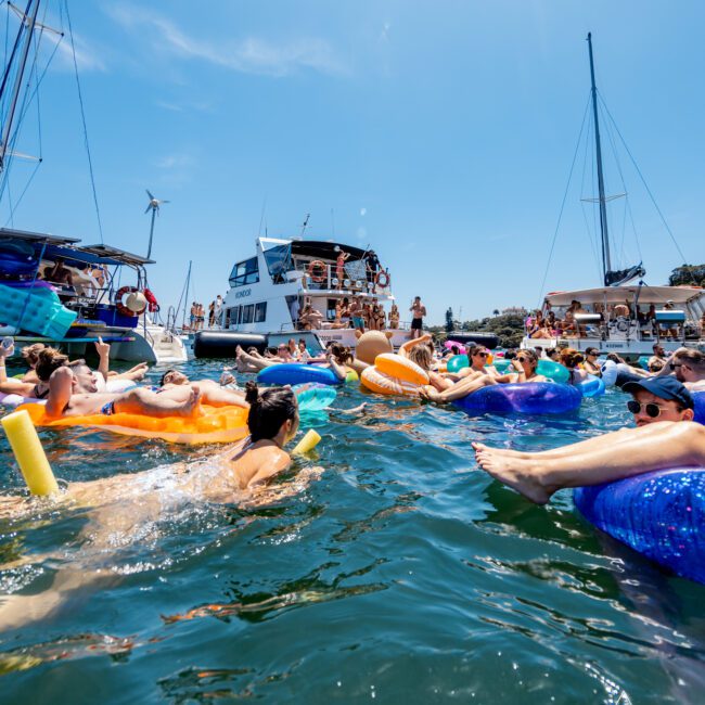 People relaxing on colorful pool floats in the sea near several anchored yachts on a sunny day. The sky is clear and blue, and the scene is lively with people enjoying the water.