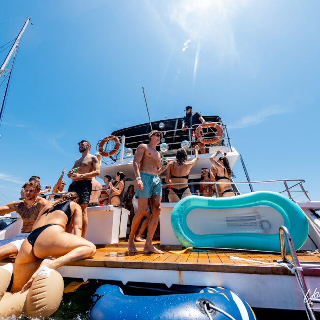 A group of people enjoys a sunny day on a yacht. Some are swimming or lounging in the water with inflatable toys, while others relax on the deck. The sky is clear and the atmosphere is lively and festive.