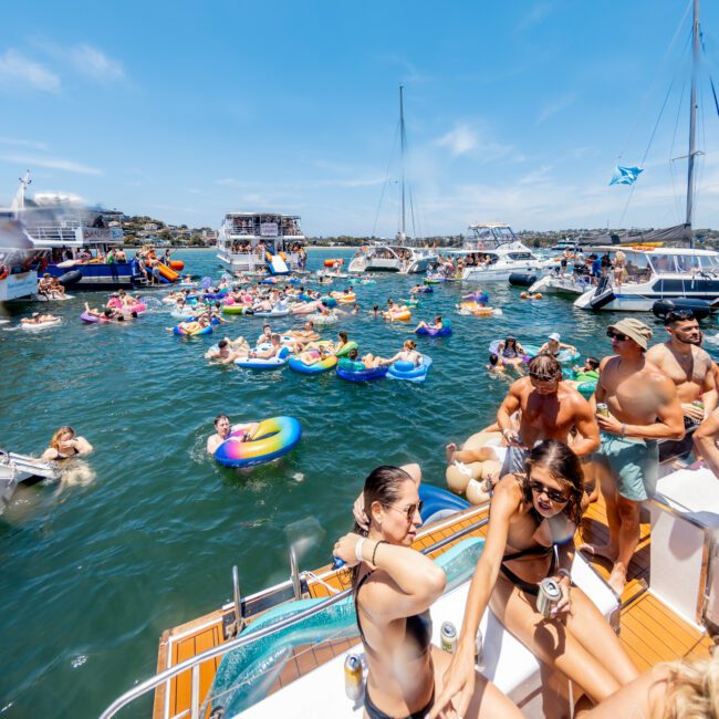 A lively scene on a sunny day with people enjoying a boat party. Several people are in swimsuits, lounging on inflatables, or standing on boats. The water is filled with yachts and colorful floats, creating a festive atmosphere.