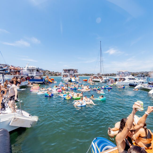 A lively scene of people enjoying a boat party on a sunny day. Numerous boats float on the water, with many individuals relaxing on colorful inflatables. People are dancing, swimming, and socializing under a clear blue sky.