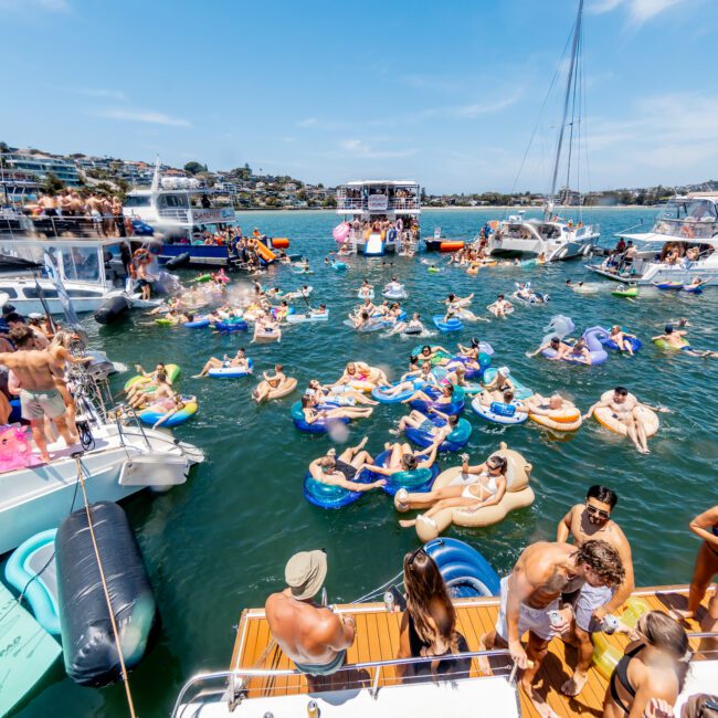 A lively boat party on a sunny day with people on boats and colorful inflatables in the water. The scene includes yachts and houses visible in the background, capturing a vibrant summer gathering.