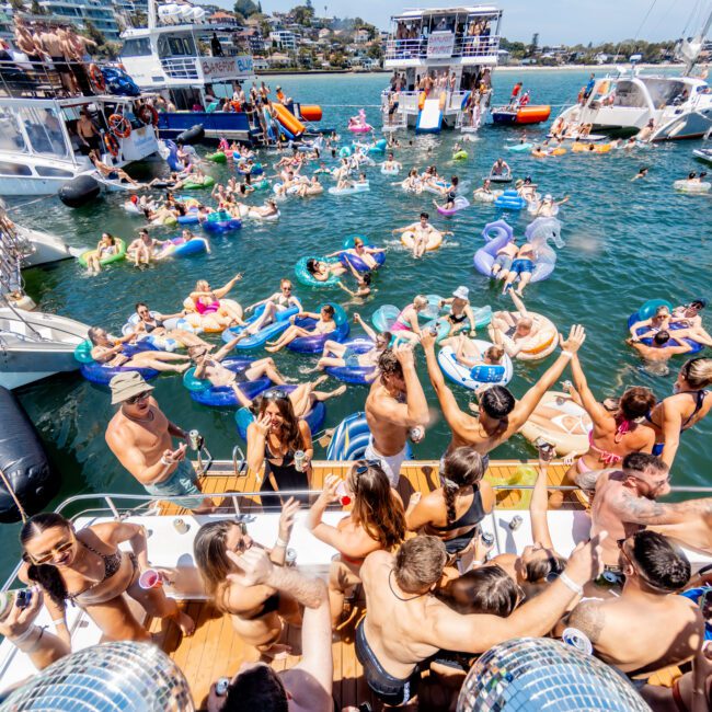 A lively scene featuring a large group of people enjoying a sunny day on a lake. Many are on inflatable floats and in the water, surrounded by boats. Others are socializing on deck, with music and drinks in the vibrant atmosphere.