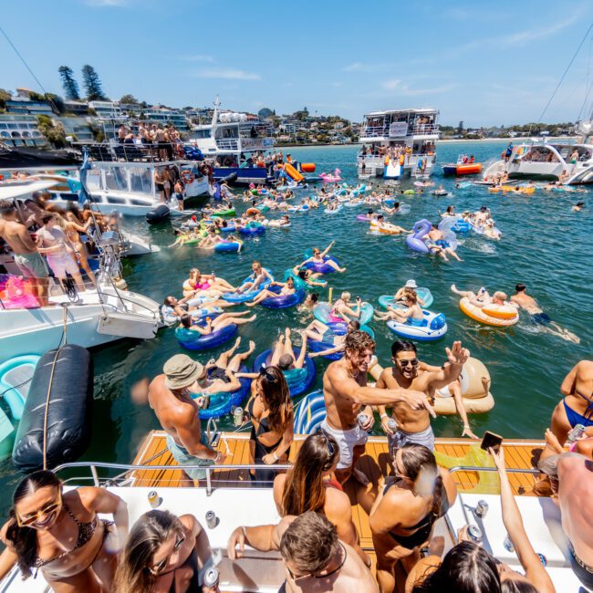 A vibrant scene of people enjoying a sunny day on the water. Numerous individuals are on boats and inflatable floats, socializing and relaxing. The backdrop features a marina with several yachts and buildings along the shoreline.