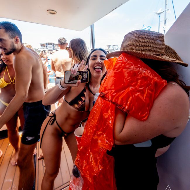 A group of people are enjoying a sunny day on a boat. A woman in a bikini poses playfully, holding a phone. Another person, partially covered by an orange inflatable ring, stands beside her wearing a straw hat. The boat is docked with a marina in the background.