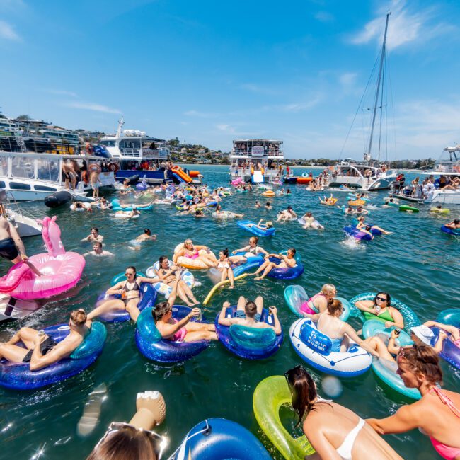 A lively scene on the water shows people relaxing on inflatable tubes and floats. Boats are anchored nearby in bright sunlight, with greenery in the background under a clear blue sky.