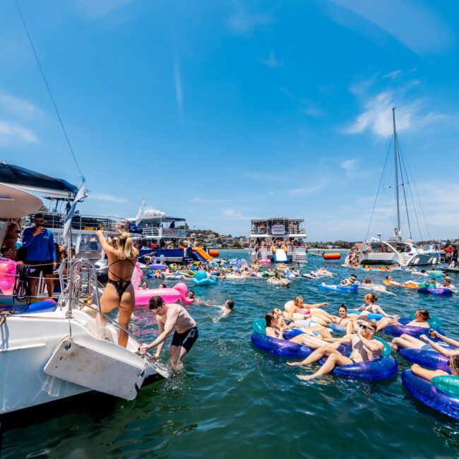 A lively scene on the water with people enjoying a sunny day. Many are on inflatable floats and boats, swimming and socializing. Others engage in activities on various vessels nearby under a clear blue sky.