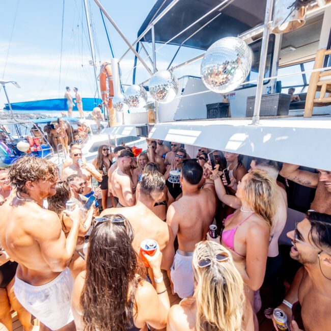 A lively group of people in swimsuits enjoy a yacht party under sunny skies. They are gathered on a deck, surrounded by disco balls and boats. Some hold drinks, while others chat and laugh, capturing a festive and carefree summer atmosphere.