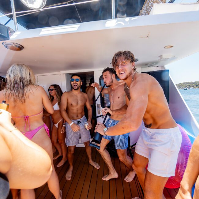 A group of people in swimwear are enjoying themselves on a yacht deck. Some are dancing while others smile at the camera. The sun is shining, and there are reflections of bright lights above them.