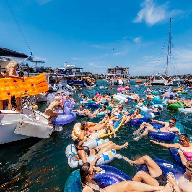 A lively scene of people floating on inflatable tubes and rafts in clear blue water, surrounded by boats. The sunny day adds to the festive atmosphere as everyone enjoys the water and socializes.