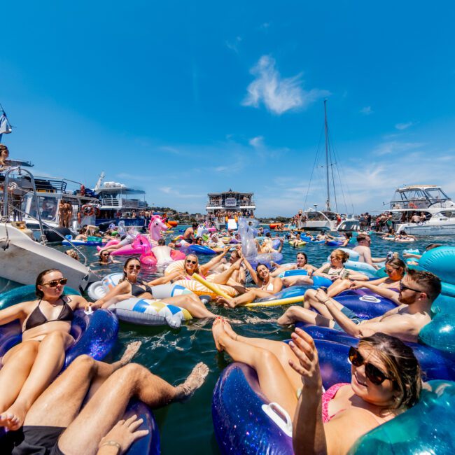 A large group of people enjoying a sunny day on the water, floating on colorful inflatables surrounded by boats. The scene shows a festive atmosphere with clear blue skies and vibrant summer fun.