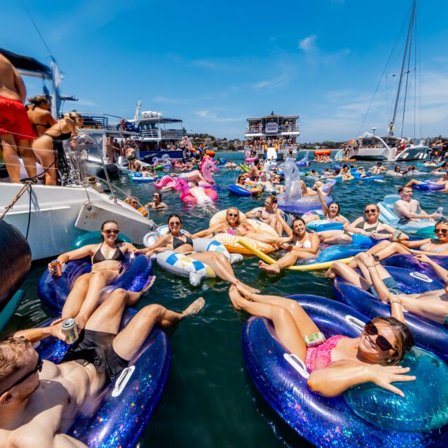 A lively gathering of people in swimsuits floats on inflatable tubes in a sunny, crowded marina. Boats are anchored nearby, and revelers are relaxing and socializing on the water. The atmosphere is festive and vibrant.