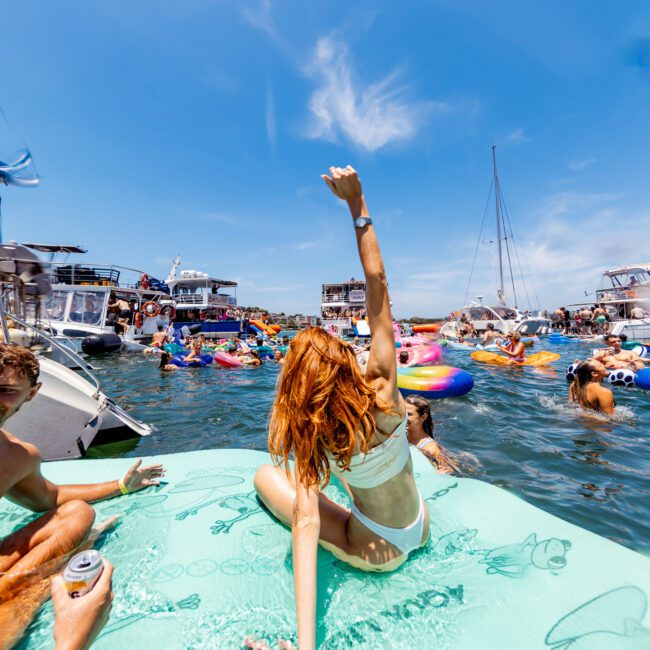 A lively scene of people enjoying a sunny day on a lake. They are on boats, floating on inflatables, and swimming. A woman in a swimsuit sits on a giant float with her hand raised. The sky is clear and blue.