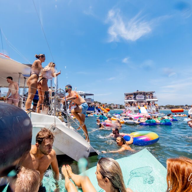 A lively scene of people enjoying a sunny day on boats and inflatable floats. A group is gathered on a catamaran, while others swim and relax nearby. The water is filled with colorful floats, and the sky is clear and blue.