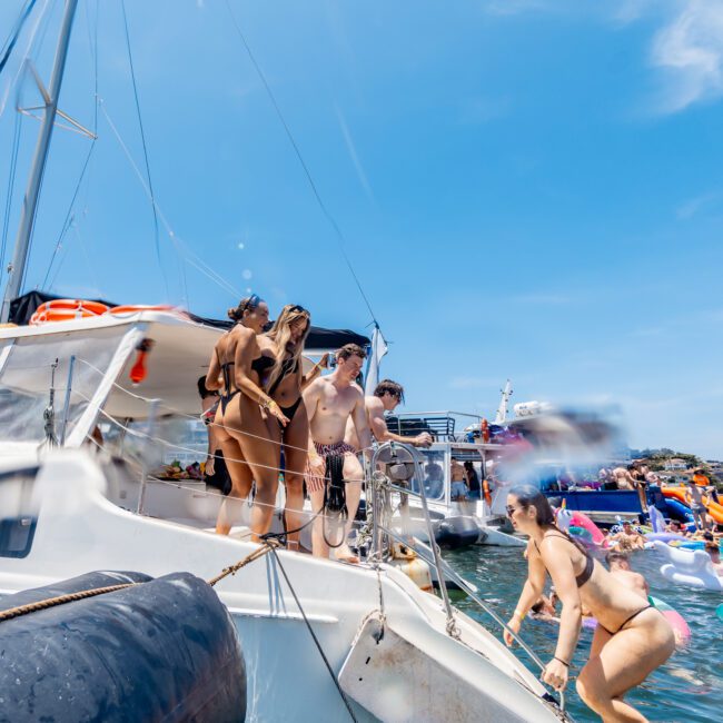 A group of people in swimsuits stand on a boat deck, enjoying a sunny day on the water. Some are entering the water by a small ramp. There are other boats and inflatables in the background, under a clear blue sky.