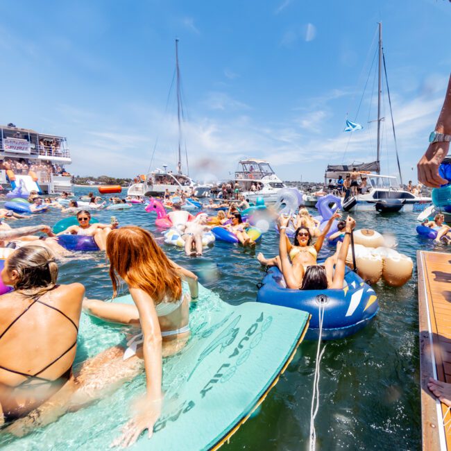 A vibrant scene of people enjoying a sunny day on the water, floating on inflatable rafts and toys. Boats are anchored nearby and individuals are relaxing and socializing. The sky is clear and blue, enhancing the festive atmosphere.