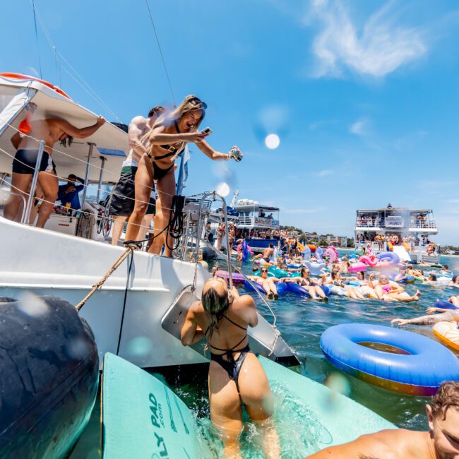 Two people on a boat prepare to join a lively lake party. One is sliding a large float into the water, while another pours a drink. The lake is filled with people on colorful inflatables, and the sky is clear and sunny.