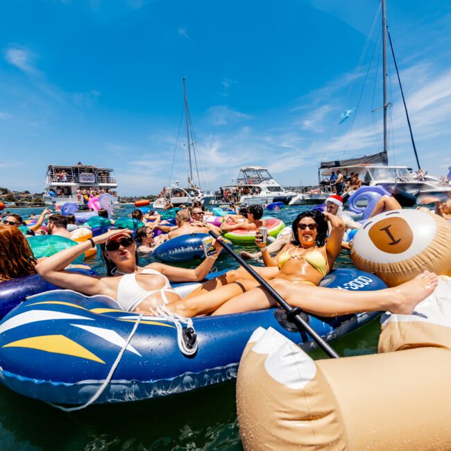 People lounging on inflatable rafts and floats in a crowded body of water, with yachts in the background. The vibrant scene features blue skies and is filled with summer-themed inflatables, creating a festive and relaxed atmosphere.