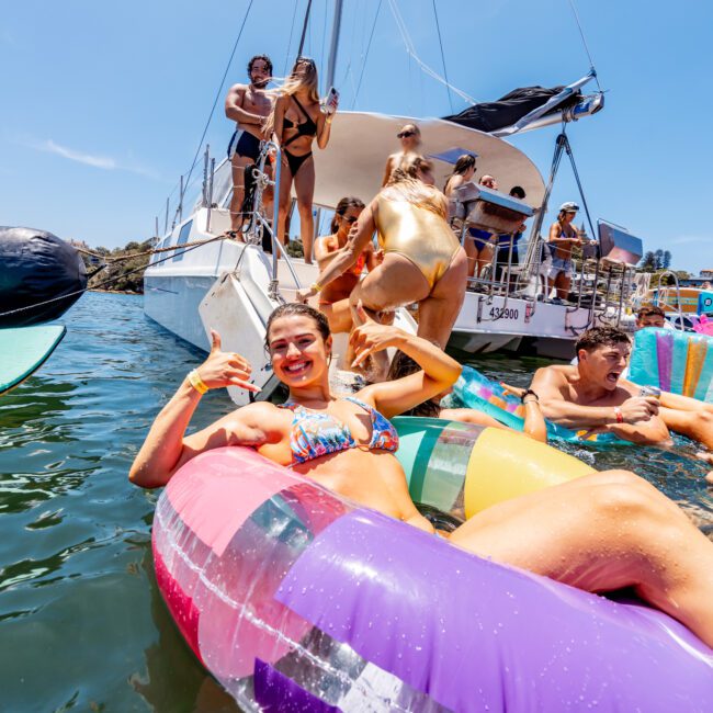 A group of people enjoys a sunny day on the water, lounging on colorful inflatables. A boat is in the background, with some people on board. The atmosphere is lively and relaxed.