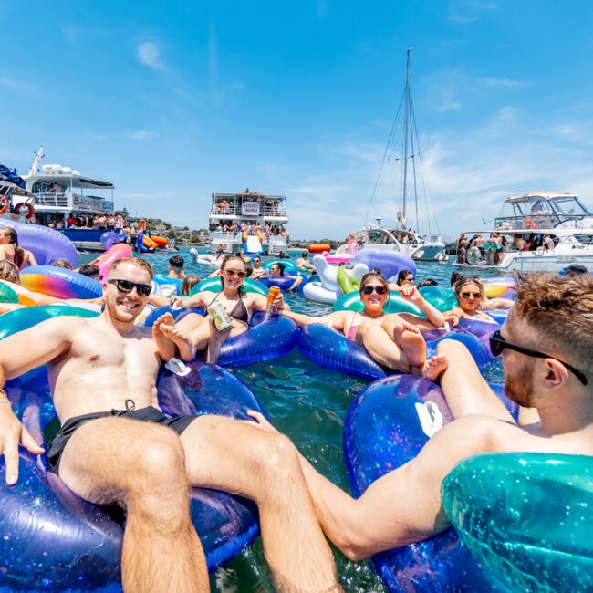 A group of people relaxing on inflatable floats in a sunny, blue-water marina, surrounded by boats and yachts. The sky is clear, and everyone appears to be enjoying a lively outdoor gathering.