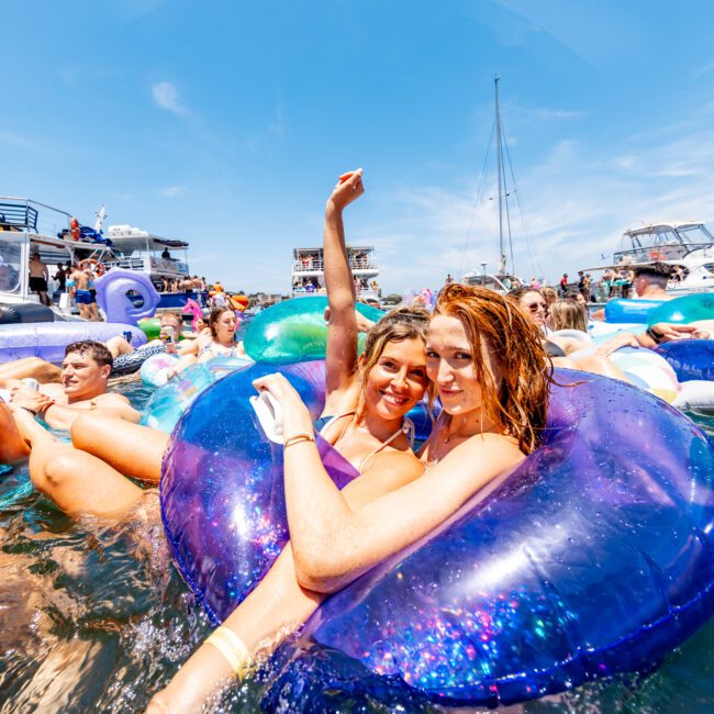 People are enjoying a sunny day on the water, lounging on colorful inflatable tubes. Boats are anchored nearby in the background. Two women smile and pose for a photo in the center, surrounded by others having fun.