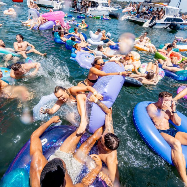 A lively scene of people enjoying a party on a sunny day, floating on large inflatable rafts in the water. Boats are anchored nearby. The crowd is diverse, with many colorful inflatables creating a festive atmosphere on the water.