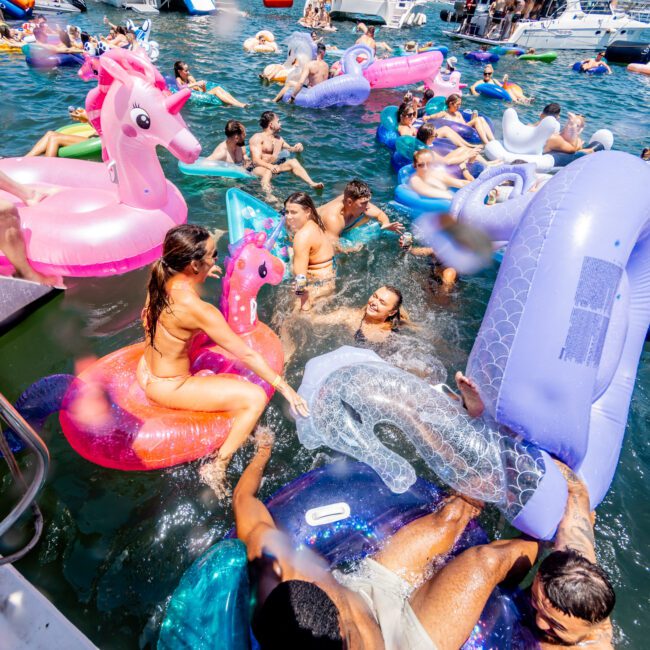 A vibrant scene of people enjoying a sunny day on the water, floating on colorful inflatable rafts, including unicorns and other fun shapes. Several boats are anchored nearby, with people socializing and relaxing.