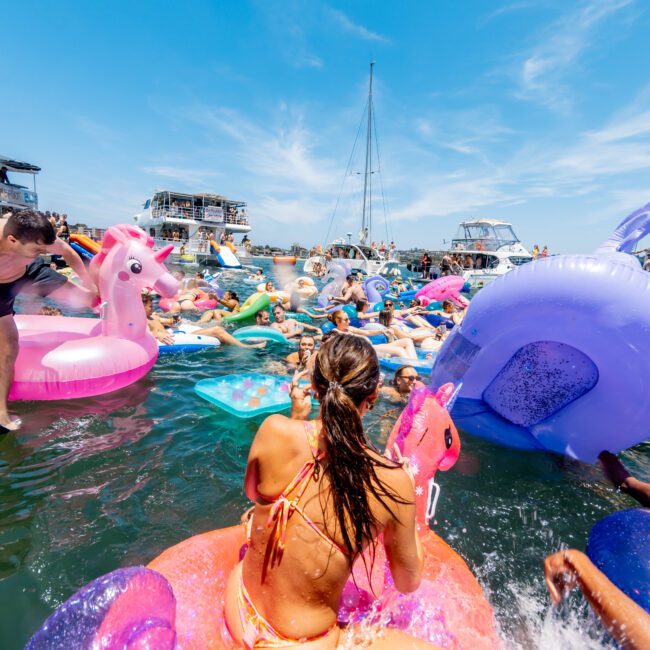 A crowded summer scene on a lake with people lounging on colorful inflatable floats, including a pink flamingo and unicorn. Boats and a sailboat are anchored nearby under a clear blue sky.