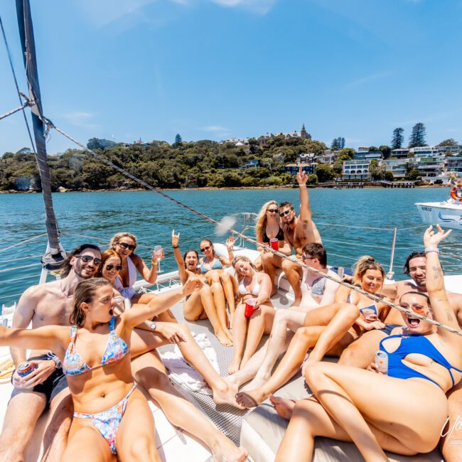 A group of cheerful people relaxes on a boat deck, enjoying sunny weather. They are smiling and making peace signs, surrounded by clear blue water and distant tree-covered hills. The scene is lively and joyful.