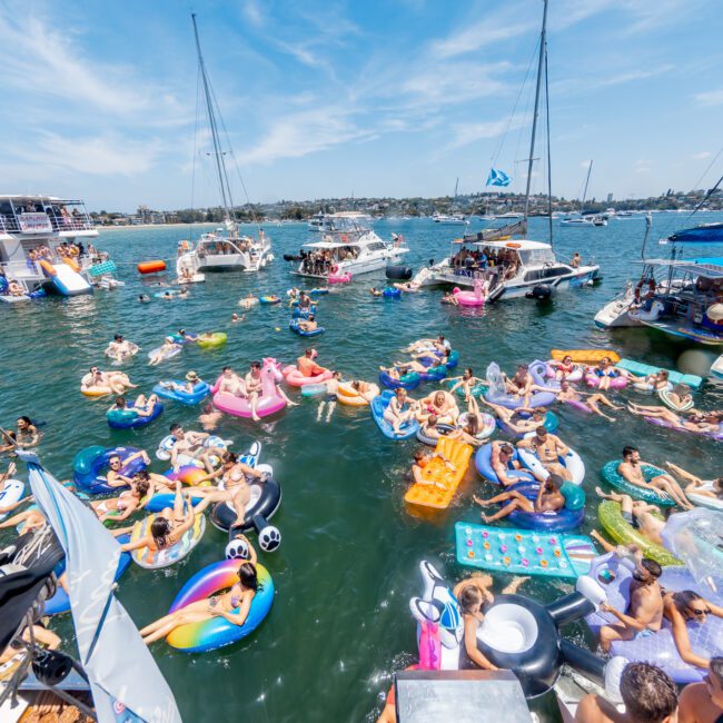 A lively scene on a sunny day, with numerous people relaxing in colorful inflatable tubes and floats on the water. Boats are anchored around the area, and people are enjoying the festive atmosphere under a clear blue sky.