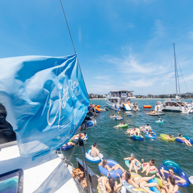 A lively scene of people relaxing on inflatable floats in a sunny bay, surrounded by boats. A blue flag with white writing flutters in the foreground, capturing the festive and carefree atmosphere.