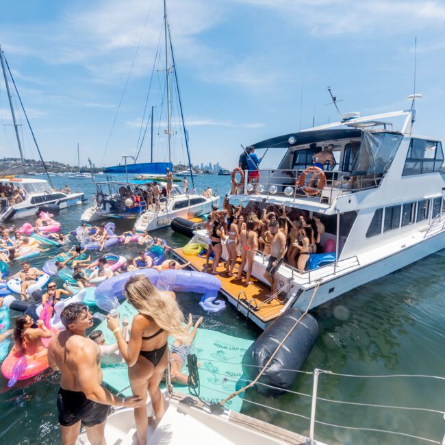 A lively boat party on a sunny day with people on a large yacht and others floating on inflatable pool toys in the water. The scene is set against a backdrop of sailboats and a clear blue sky.