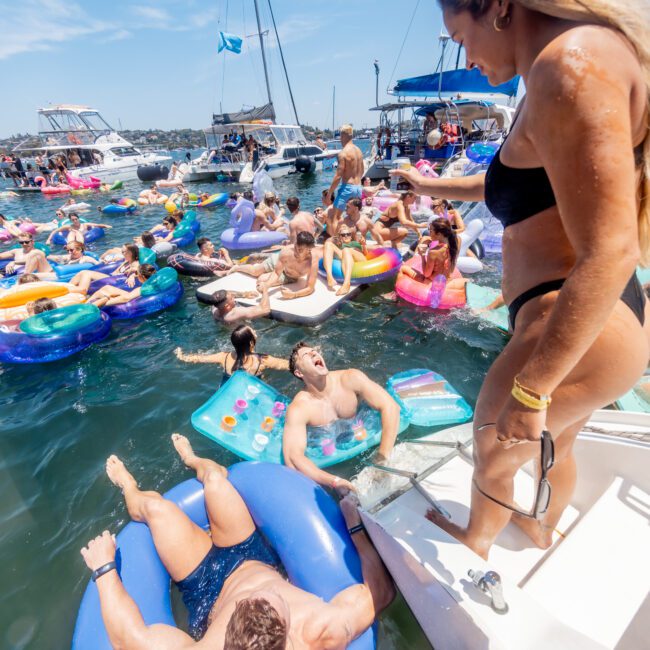 A lively scene on a sunny day with people enjoying a boat party. Individuals in swimwear are on various colorful inflatables in the water. A woman in a black bikini stands on a boat, while others relax and socialize in the background.