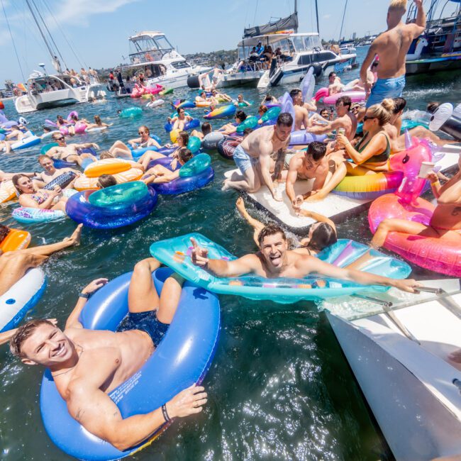 A lively scene of people enjoying a sunny day on floaties in the water near several boats. The group appears relaxed and cheerful, with vibrant inflatable rings and a variety of water inflatables enhancing the festive atmosphere.